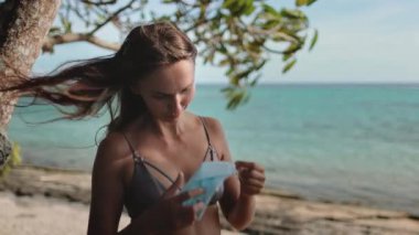 Güneşli bir sahilde genç bir kadın, dikkatlice koruyucu bir yüz maskesi takıyor, yeni seyahat normalinin farkındalığını ve bakımını somutlaştırıyor