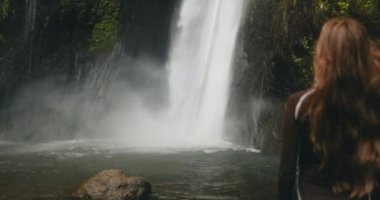 Güçlü bir orman şelalesinin yanında duran turist kız. Uzun saçlar rüzgarda savruluyor. Turizm, yürüyüş, yürüyüş, rahatlama, aktif yaşam tarzı, Bali 'de yaz tatili. Yavaş çekim 4K arka görünüm yakınlaştır