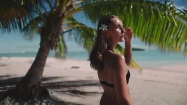 Genç bir kadın kumsalda güneşli bir günün tadını çıkarıyor, saçlarında bir çiçekle bir palmiye ağacının altında duruyor, güneşin sıcaklığını ve doğanın güzelliğini hissediyor.