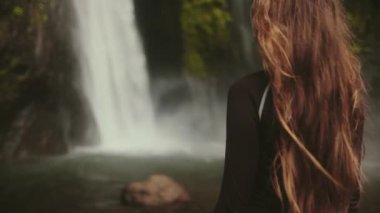 İnsanlar ağır çekim orman şelalesi. Uzun saçlı kadın, egzotik yağmur ormanlarındaki yüksek şelaleli tropik ada manzarasının tadını çıkarıyor. Bali Endonezya. Yaz tatilindeki bir kadın. Arka planı kapat