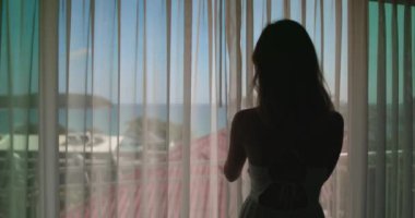 Pencere kenarında durup, tropik bir Tayland tatil köyünün engin okyanusuna bakan bir kadın..