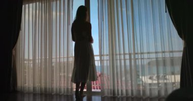 Genç bir kadın otel odasındaki perdeleri açarken, tropik bir sahile bakan parlak bir pencerenin önünde siluete bürünür. Bu anımsatıcı görüntüyle seyahat ve tatilin güzelliğinin tadını çıkar.