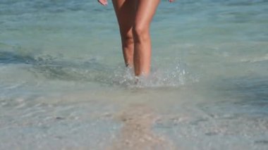 Kadın bacakları deniz suyundan çıkar yavaş çekimde kapanır. İnce bronzlaşmış kadın ayakları kristal su dalgaları sıçratıyor. Kum sahili kıyısı. Yaz tatilindeki turist. Yaşam tarzı tatil konsepti