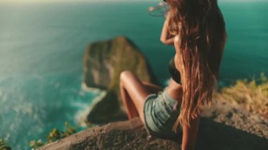 Kadın dağ yamacında oturup günbatımının tadını çıkarıyor. Rahatla, özgürlük, ilham. Rüzgar uzun saçlar savuruyor. Bali yaz tatilindeki kadın turist. Sinematik çekim, kamera yükseliyor, yavaş çekim