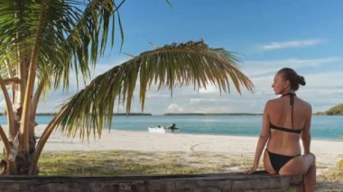 Genç bir kadın palmiye ağacının altındaki ahşap iskelede oturuyor. Fransız Polinezyasındaki güzel bir gölün tropikal manzarasının tadını çıkarıyor.