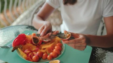 Bir tabak tropikal meyve, kadın el emeği, kaşıkla tutku meyvesi yiyor. Beyaz bir tabakta çeşitli egzotik meyveler. Tropik yaz tatilinde sağlıklı yiyecekler yemek, sağlıklı atıştırmalıklar..
