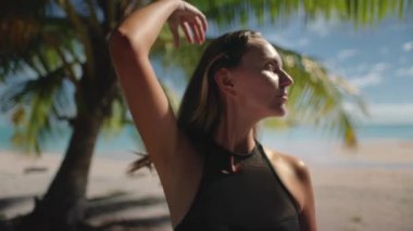Genç bir kadın, tropik bir sahilde güneşin ve esintinin tadını çıkarıyor. Arkasında turkuaz bir deniz ve bir palmiye ağacı var.