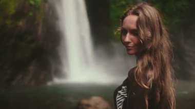 Güçlü Bali şelalesinin önünde, yemyeşil bir şelaleyle çevrili bir kadın. Su aşağı akarak dingin bir an için dramatik bir zemin oluşturur. Air Terjun Munduk, Eriyor. Yavaş çekim