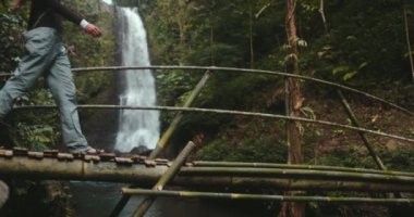 Yürüyüşçü kız Bali adasında orman şelalesinin tadını çıkarıyor. Uzun saçlı turist kadın bambu köprüsünde yürüyor ve güçlü şelalenin keyfini çıkarıyor. Seyahat tatili. Eriyen Şelaleler. Yavaş çekim