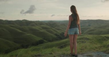 Yeşil tepedeki çayırda duran kadın, ellerini kaldırdı, rüzgar saçlarını süpürdü - özgürlük, doğanın tadını çıkarın. Turist kız gün batımını seyrediyor. Güzel manzara. Açık hava yaşam tarzı yaz tatili tatili. Yavaş çekim