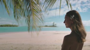 Bir kadın bir plajın tepesinde, bir palmiye ağacının yanında, denizin ve güneşli havanın tadını çıkarıyor..
