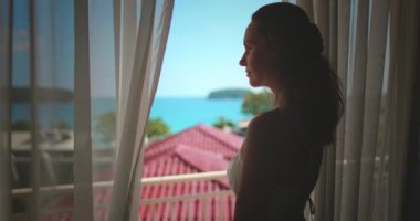 Güneşli bir günde pencerenin yanında durup okyanusa bakan bir kadın. Tropikal bir ülkedeki otel odasının manzarasının tadını çıkarıyor.