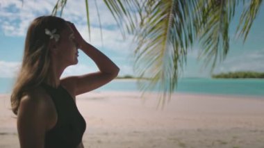 Bir kadın kumlu bir sahilde, güneşli bir günde okyanusa bakan yüksek bir palmiye ağacının yanında duruyor..