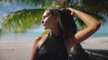 Bir kadın kumlu bir sahilde, açık mavi bir gökyüzünün altında, arka planda hafif dalgalarla uzun bir palmiye ağacının yanında duruyor..