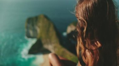 Kadın saçı rüzgarda ağır çekimde savruluyor. Okyanus manzaralı uçurumun kenarında oturan turist kızın arkası. Seyahat, turizm, tatil. Endonezya, Nusa Penida Adası, Kelingking Gizli Nokta Sahili
