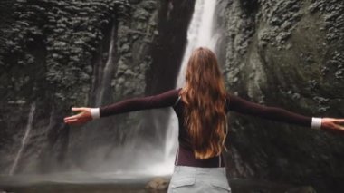 Dramatik orman tropikal şelalesi. Turist kadın muhteşem vahşi doğa manzarasına bakmak için ellerini kaldırdı. Seyahat, turizm, özgürlük, aktif yaşam tarzı. Bali Adası, Terjun Munduk Havayolları. Yavaş çekim