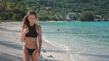 Genç bir kadın, kristal berrak turkuaz su ve canlı yeşillik ile çevrili tropikal bir plajda geziniyor, cennetteki kaygısız yaz titreşimlerini somutlaştırıyor.