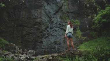 Turist, tropikal bir ormandaki uçurumdan akan bir şelale manzarasının keyfini çıkarıyor. Bir kayanın üzerinde duruyor, doğanın güzelliğine hayran.