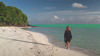 Kadın güneşli bir günün tadını çıkarıyor. Turkuaz su ve yemyeşil bitki örtüsüyle tropik bir kumsalın beyaz kumlarında yürüyor.