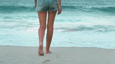 Ağır çekimde kadın ayakları kumlu kumsalda çıplak ayakla yürüyor ve kumsalda ayak izleri bırakıyor. Okyanus dalgaları, yaz tatilindeki ince kadın turistin arka görüntüsü. Seyahat, turizm, tatil, dinlenme, aktif yaşam tarzı