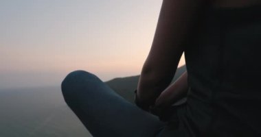 Dağın tepesinde oturan kadın, karanlık dramatik günbatımı çekimi. Okyanus koyuna ve dağ manzarasına bakan turist kız portresi. Seyahat, turizm, yürüyüş, dinlenme. Kamera yukarı, yavaş çekim 4K