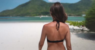 Kumlu bir plajda duran bikinili bir kadın, dalgalar yavaşça kıyıya vuruyor. Güneş mavi gökyüzünde parlıyor..