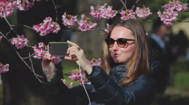 Kiraz çiçeklerinin güzelliğini yakalayan genç bir kadın, baharda Japon parkında fotoğraf ve sosyal medya aracılığıyla deneyimlerini paylaşarak neşeyle fotoğraf çekiyor.