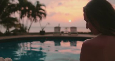 Gün batımında kız silueti yüzme havuzunun yanında. Durgun suya yansıyan renkli gündoğumu gökyüzü. Rahatla, geri çekil, meditasyon yap. Seyahat, turizm, tatil spa tatili. Yavaş çekim portresi yakınlaştırır