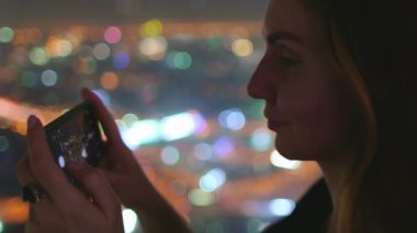 Kadın cep telefonuyla gece şehir panoramasını çekiyor. Şehir merkezindeki renkli şehir merkezi, Baiyoke Tower Sky, Bangkok, Tayland 'ın en üst katının gece lambaları manzarası. Yaklaşın, ağır çekim.