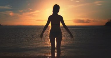 Tropik bir plajda kollarını açmış okyanusun üzerinde günbatımının tadını çıkaran genç bir kadının silueti. Güneş ufukta batıyor, suyun üzerine sıcak bir parıltı saçıyor.