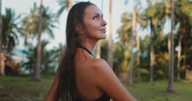 Bikinili bir kadın Tayland 'da yemyeşil palmiye ağaçlarının önünde güven içinde duruyor. Sahne yavaş çekimde, tropikal ortamın güzelliğini vurguluyor..