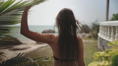 Egzotik bir adada tropikal palmiye bahçesinde yürüyen bir kadın. Bikinili bir kız lüks otelin okyanus manzarasının tadını çıkarıyor. Seyahat, turizm, tatil, tatil. Arka plan, yavaş çekim