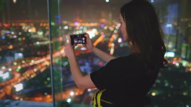 Turist kadın gözlem güvertesinde duran cep telefonuyla şehir manzarası manzaralı panoramasını çekiyor. Kız akıllı telefondan şehir ışıklarının fotoğraflarını çekiyor. İş merkezinin en üst katı, kule manzaralı.