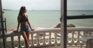 Güzel kız yaz tatilinde tatil köşkünün terasında dinleniyor. Arka planda tropik ada deniz manzarası boş kumsal sahili. Tayland egzotik adasına seyahat.