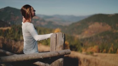 Kırsal bir çitte oturan bir kadın, gözleri uzakta beliren görkemli dağlara sabitlenmiş. Düşüncelere dalmış gibi, sakin doğayla çevrili..