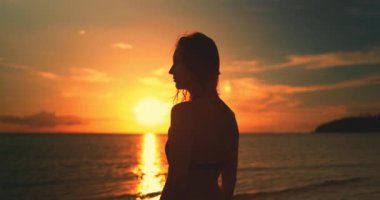 Kadın sahilde durup Tayland 'da okyanusun üzerinde güzel bir günbatımını seyrediyor. Gökyüzü turuncu ve altındır, suyu yansıtır.