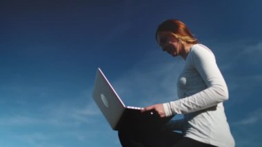 Modern giysiler içindeki genç bir kadın dizüstü bilgisayarına gömülmüş kırsal ahşap bir çitte zarifçe oturuyor. Güneş onun üzerine doğayı ve teknolojiyi harmanlayarak dijital dünyayı dolaşırken parlıyor..