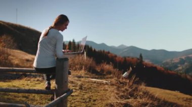 Yaratıcı kadın kırsal kesimde oturuyor, dizüstü bilgisayarla çalışıyor. Güzel kırsal manzaranın tadını çıkar. Freelancer sonbahar dağında günbatımını uzaktan takip eder. Seyahat, özgürlük, aktif yaşam tarzı, çevrimiçi dijital iş