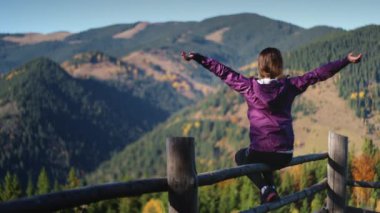 Turist kadın dağlarda sabahtan zevk alıyor. Çitin üzerinde oturan kız ellerini kaldırarak güzel dağlık araziye bakıyor. İdeal dinlenme yeri kavramı. Seyahat, turizm, tatil, yürüyüş