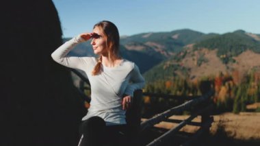 Genç bir kadın çitin üzerinde oturuyor, gözlerini güneşten koruyor ve uzaktaki dağların manzarasının tadını çıkarıyor.