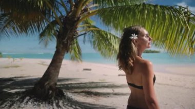 Genç bir kadın, beyaz kum ve turkuaz suyla tropikal bir sahilde dinleniyor. Güneşin ve dalgaların sesinin tadını çıkarıyor. O, saçına bir çiçek ve siyah bir bikini giyiyor.