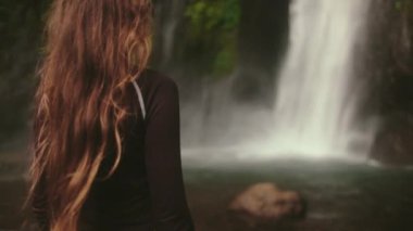 Genç bayan turist, tropikal yağmur ormanlarındaki yemyeşil bir uçurumdan dökülen görkemli bir şelale manzarasının tadını çıkarıyor.