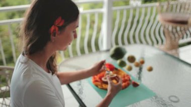 Turist, yemyeşil manzaralı bir balkonda tropikal bir kahvaltı yapıyor. Bir tabak taze papaya ve diğer egzotik meyvelerin tadını çıkarıyor, canlı ve iştah açıcı bir sahne yaratıyor.