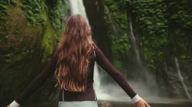 Dramatik tropik orman şelalesi manzarası yavaş çekim. Turist kız, yeşil yağmur ormanlarındaki güçlü su akıntısına karşı el kaldırıyor. Endonezya, Terjun Munduk Havayolları, Melanting, Bali Adası