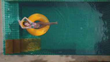 Pembe bikini giyen bir kadın Tayland 'da bir tatil köyünde sarı bir havuzda dinleniyor. O, bacaklarını açarak sırt üstü uzanıyor. Su berrak ve mavi. Hava aracı görüntüsü