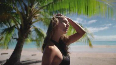 Kadın tropikal bir adada esintinin ve sıcak havanın tadını çıkarıyor. Beyaz bir kumsalda dururken saçları rüzgarda hareket ediyor.