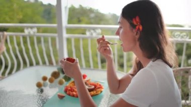 Genç bir kadın egzotik meyvelerle dolu bir balkonda oturmuş tropik kahvaltısının tadını çıkarıyor.