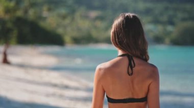 Bikini giyen bir kadın Tayland 'da bir plajda duruyor, ağır çekimde yakalanmış. Arka planda dalgalar çarptığında güneş parlıyor..