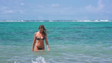 Bikini giyen bir kadın okyanus suyunda duruyor. Etrafında dalgalar çakılırken güneş onun üzerine parlıyor. Kumsalın ve suyun tadını çıkarıyor..