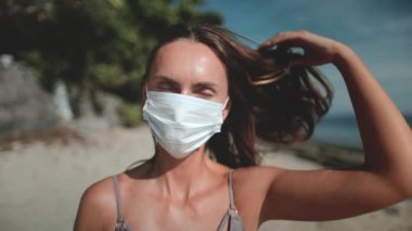 Sıhhiye maskeli kadın yaz plajında dinleniyor güneşli günün ve açık hava manzarasının tadını çıkarıyor. Bikinili kadın portresi, saçına yavaş çekimde dokunuyor. Tatilde açık hava gezisi.